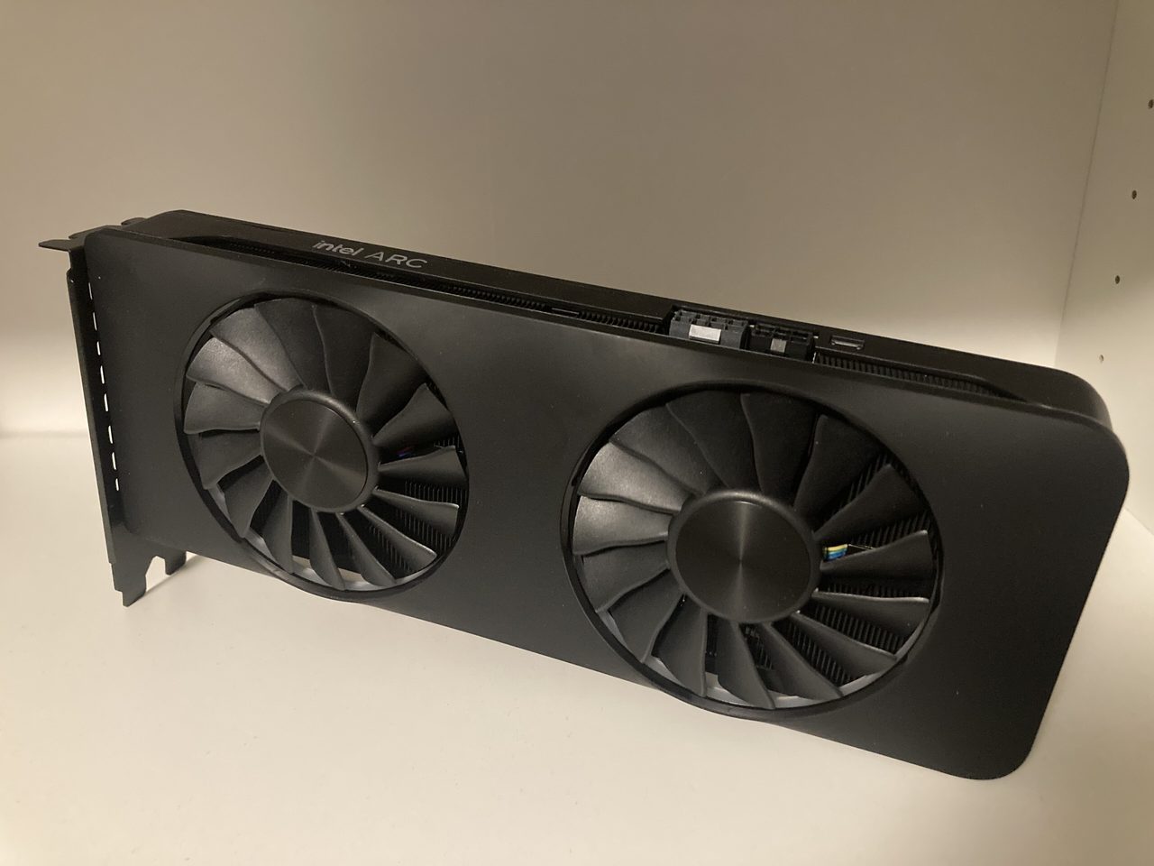 An Intel Arc A770 Limited Edition graphics card standing on a white shelf: a matt black card with two large black fans and a thin strip of lighting along its top edge, with the Intel Arc name on the side