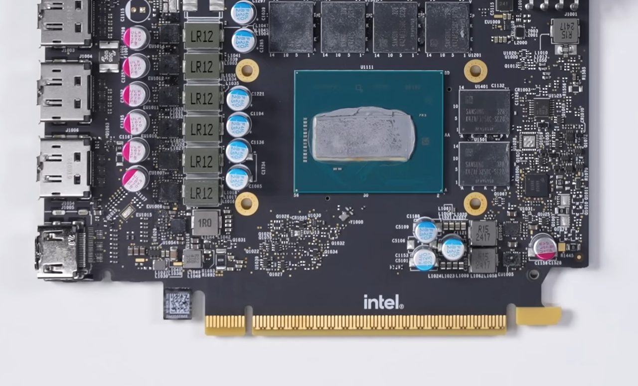 The circuit board of an Arc B580 with its cooler removed: a square GPU package with grey thermal paste in the middle, six memory chips around it, a column of power components and capacitors on the left, display connectors at the left edge and the gold PCI Express connector along the bottom with the Intel logo above it