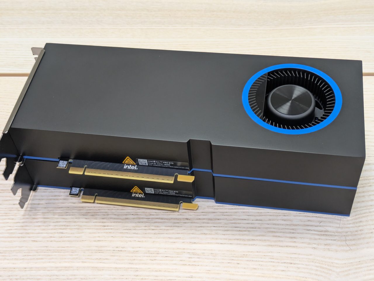 Two Intel Arc Pro B70 graphics cards stacked on a wooden surface: slim black cards with a blower fan ringed in blue at one end and a thin blue line along their sides, their gold PCI Express connectors and Intel labels facing the camera