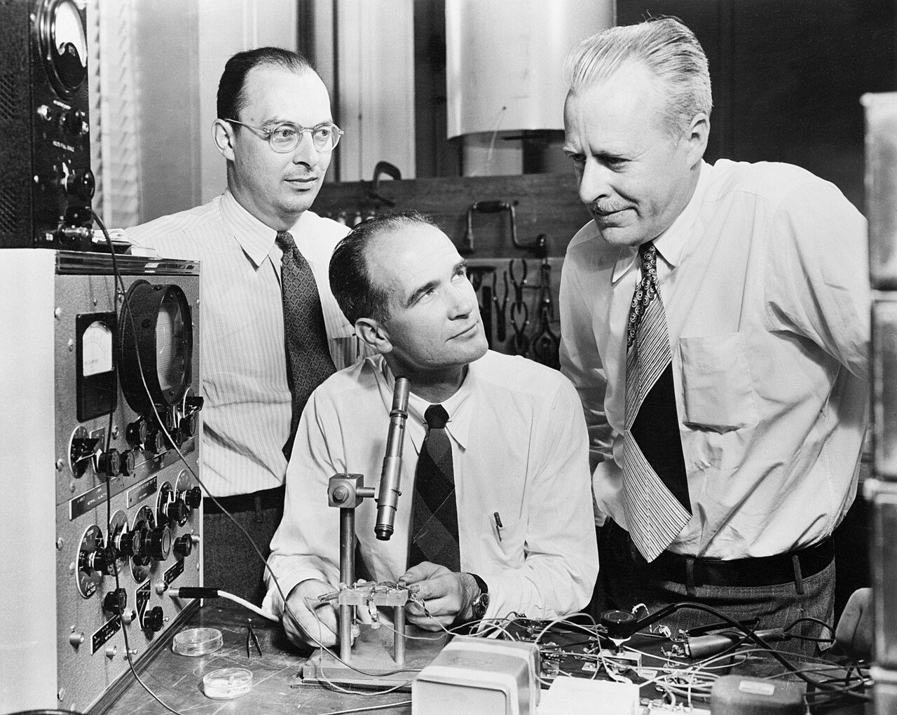 Three men in shirts and ties at a laboratory bench: John Bardeen standing on the left, William Shockley seated at a microscope in the middle, Walter Brattain standing on the right