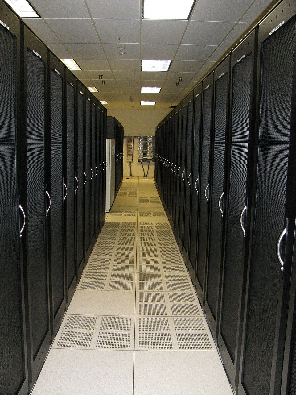 A narrow aisle between two rows of tall black server cabinets, with perforated tiles in the raised floor