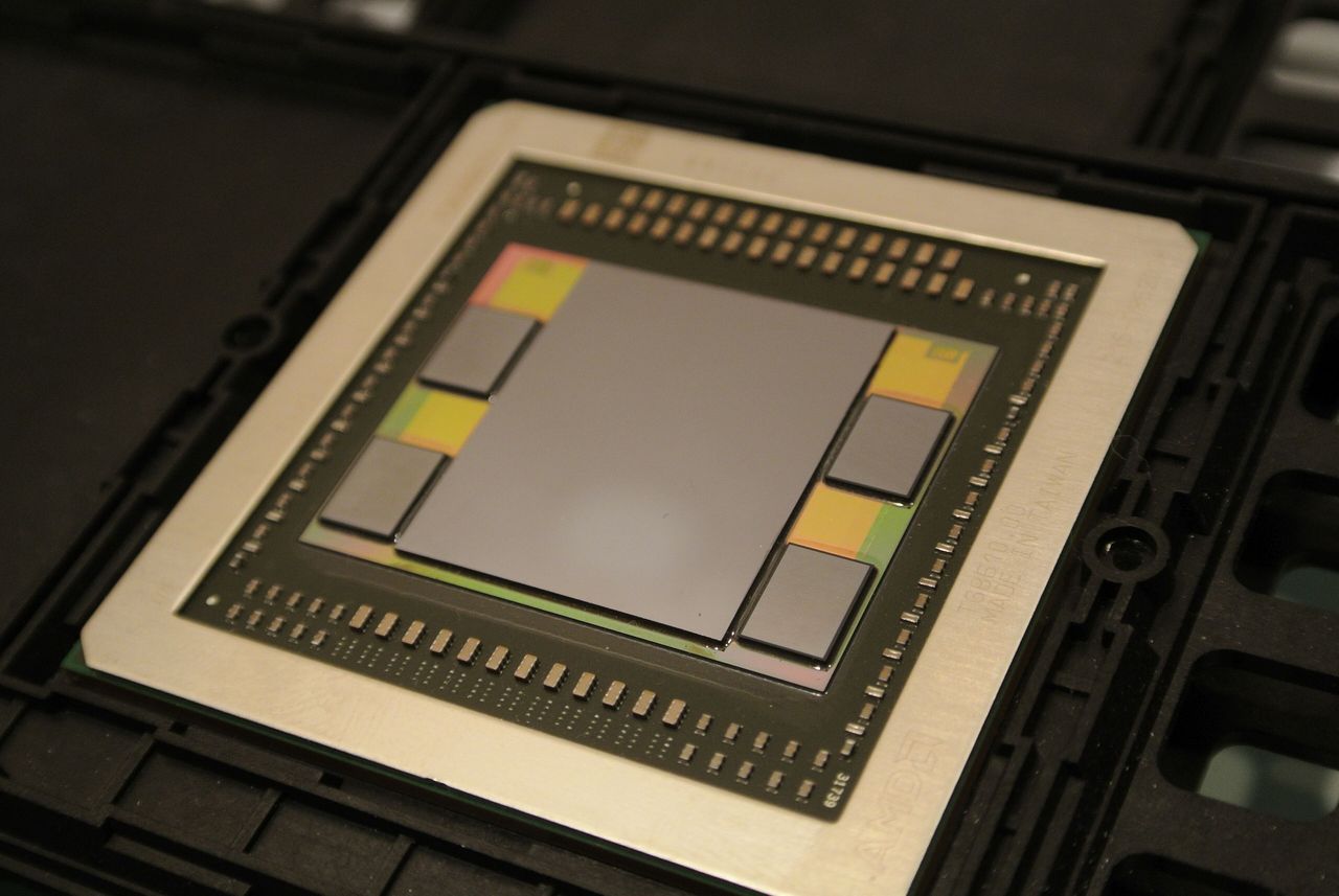 The AMD Fiji GPU package photographed at an angle: a large grey die in the centre of a metal frame, with two smaller rectangular HBM memory stacks on each side, all sitting on a silicon interposer