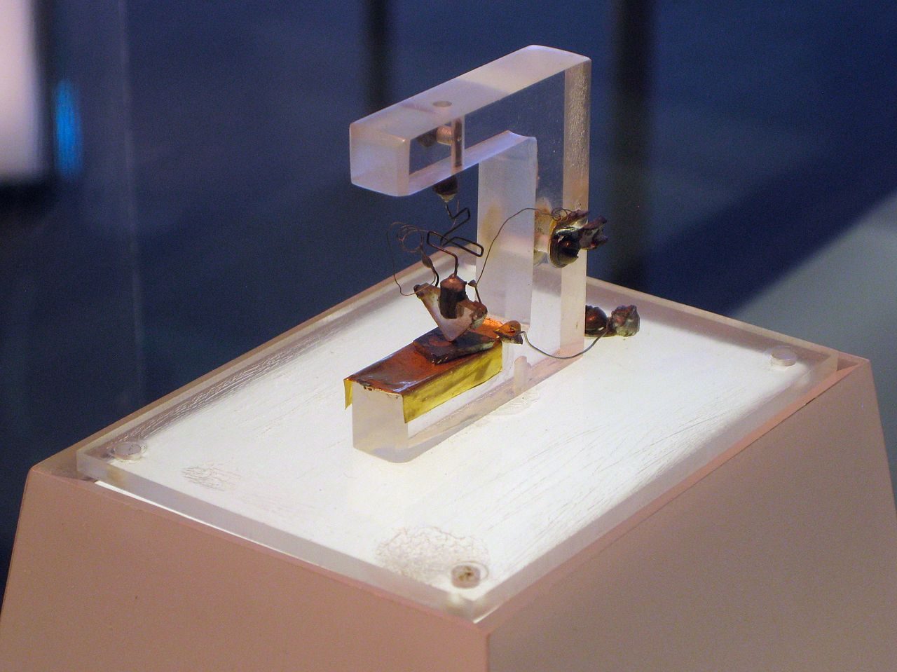 The first point-contact transistor: a clear plastic triangle held down by a bent wire spring, its tip pressed onto a small slab of germanium on a metal block, on a white plinth