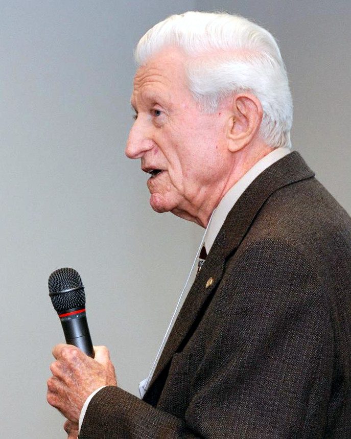 Gene Amdahl, white-haired, in a jacket and tie, speaking into a microphone