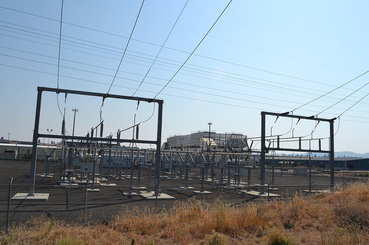 An electrical substation with steel frames and overhead power lines in the foreground and data centre buildings behind it