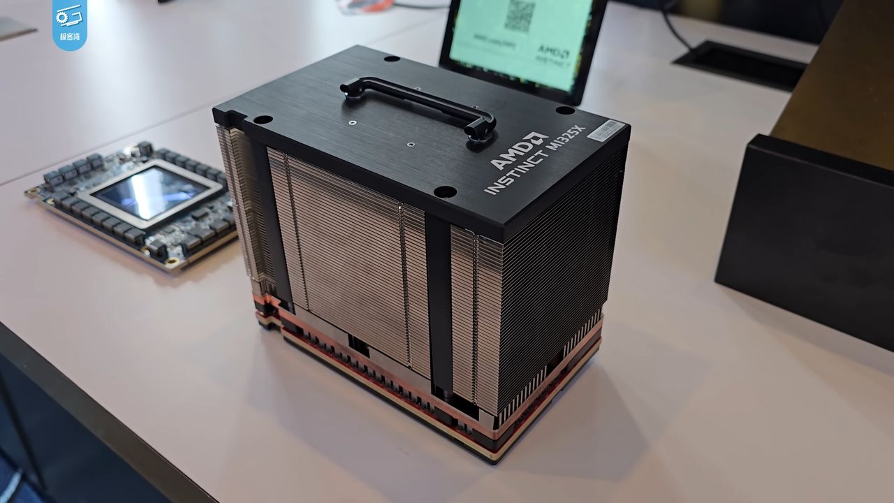 A large black and silver heatsink about the size of a shoebox on an exhibition table, with a carrying handle on top marked AMD Instinct MI325X, dense aluminium fins down its sides and copper at its base, and a bare Instinct module lying beside it