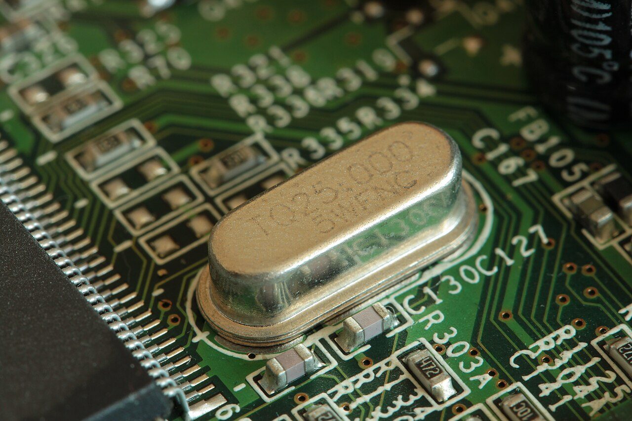A silver oval metal can marked 25.000 soldered to a green circuit board among small components