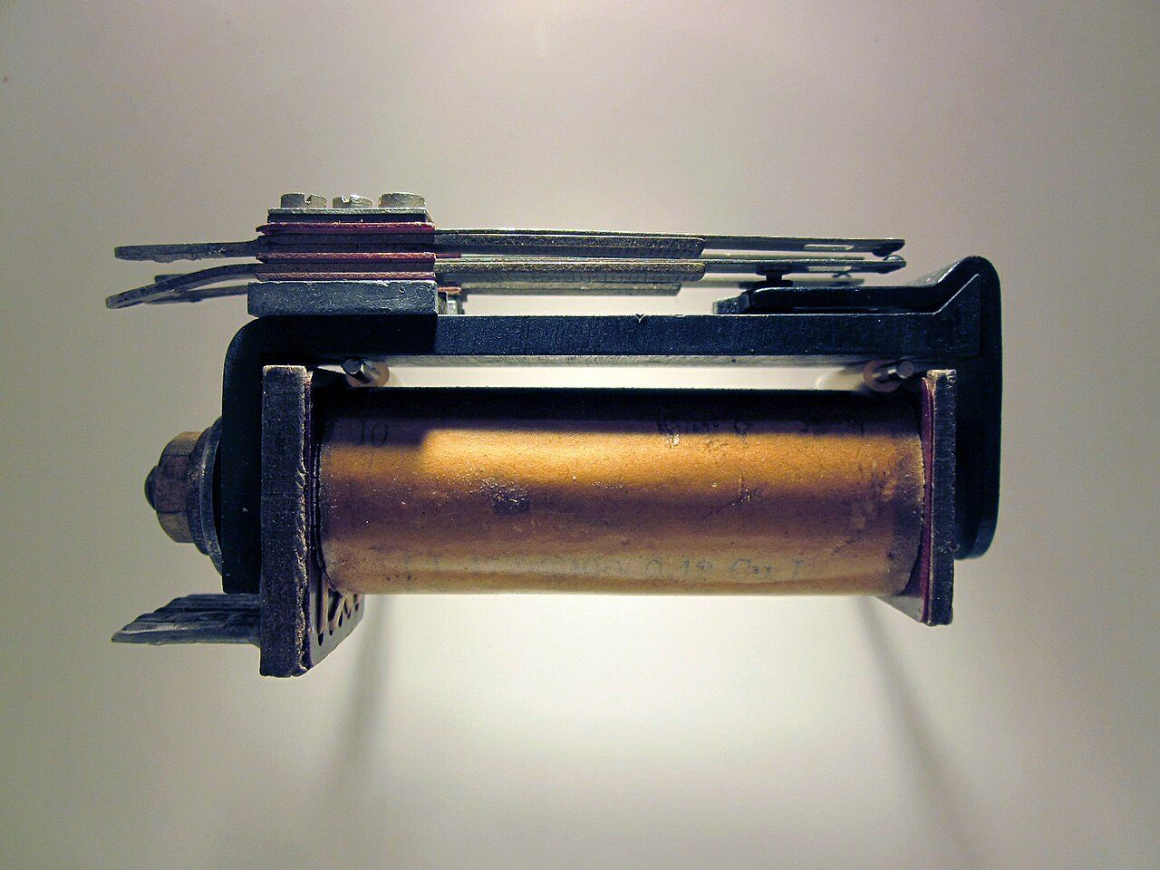 A real electromechanical relay from the 1940s seen from the side: a copper-coloured coil wound on a cylinder inside a dark iron frame, with a stack of thin metal contact strips along the top