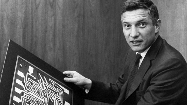 Robert Noyce in a suit holding up a large drawing of a circuit layout