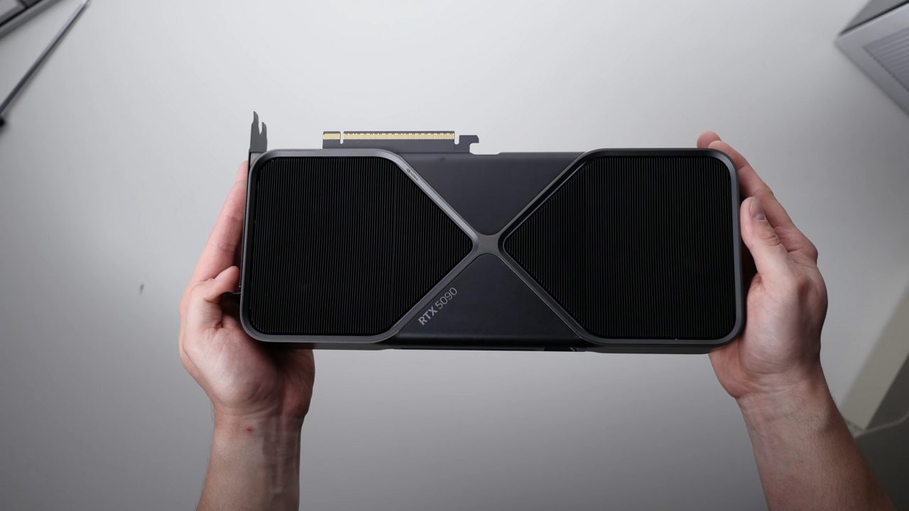 Two hands holding an RTX 5090 Founders Edition card, black with a silver X-shaped frame