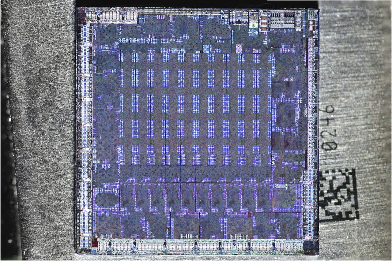An etched RV770 die photographed from above in purple and blue tones: a square of silicon covered in a regular grid of rectangular blocks, set on a grey background with a printed serial number beside it