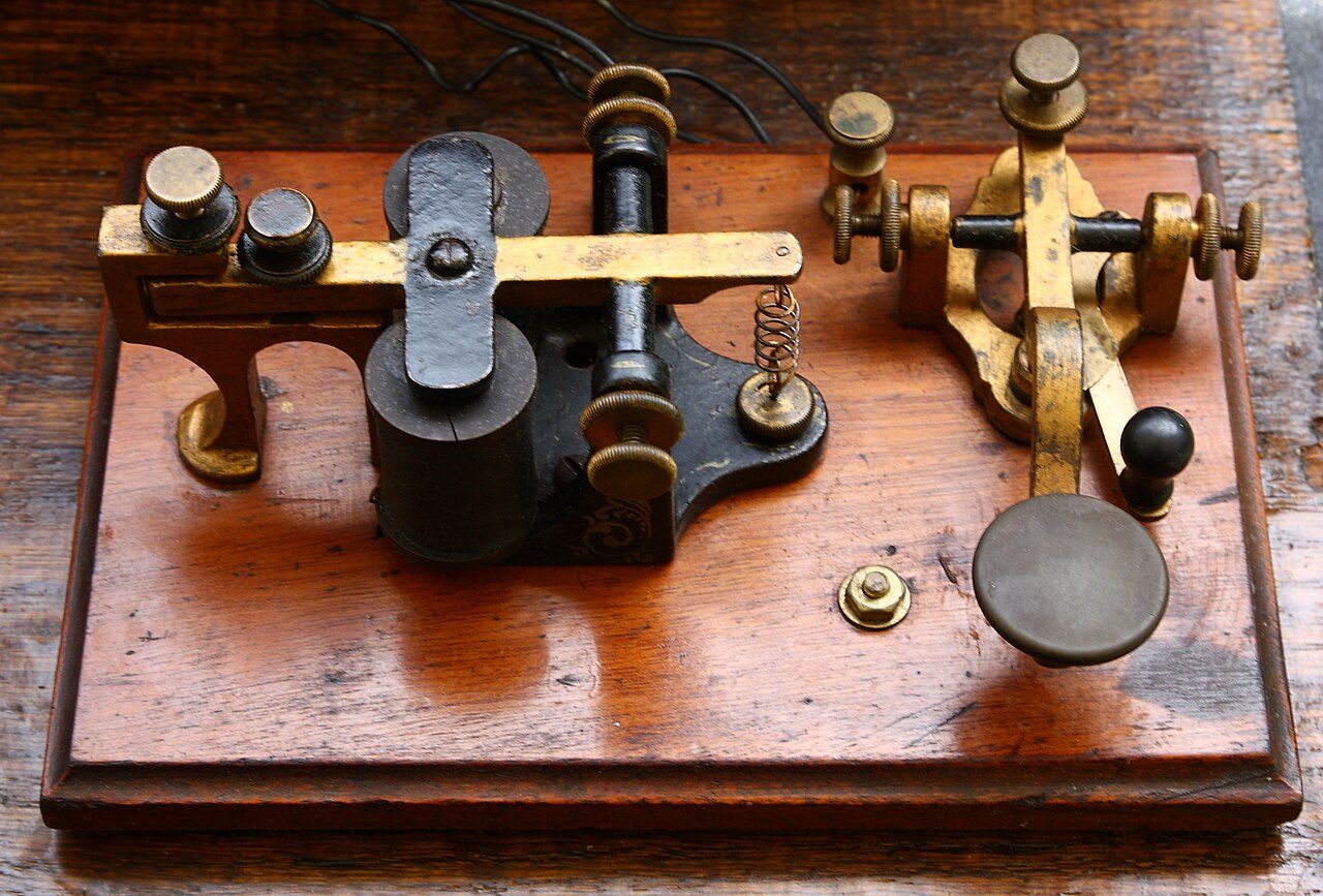 A telegraph sounder and key mounted side by side on a wooden board: on the left two dark electromagnet coils under a brass arm with a small spring, on the right a brass lever with a flat round knob
