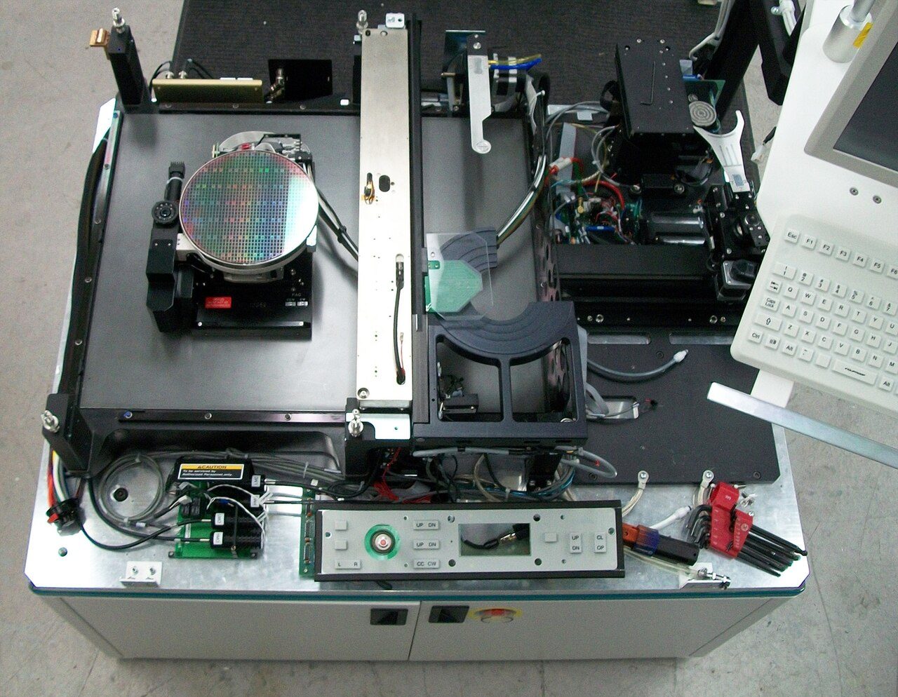 A wafer prober opened up for service: a patterned wafer on a chuck under a gantry, surrounded by motors, cables and a keyboard