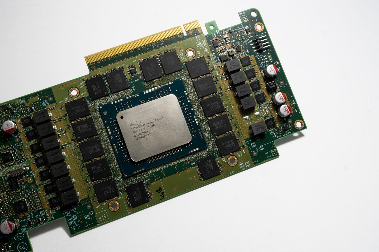 An Intel Xeon Phi 5100 coprocessor card without its heatsink: a large silver-lidded processor in the middle of a green circuit board, surrounded by a ring of black memory chips, with the gold edge connector at the top